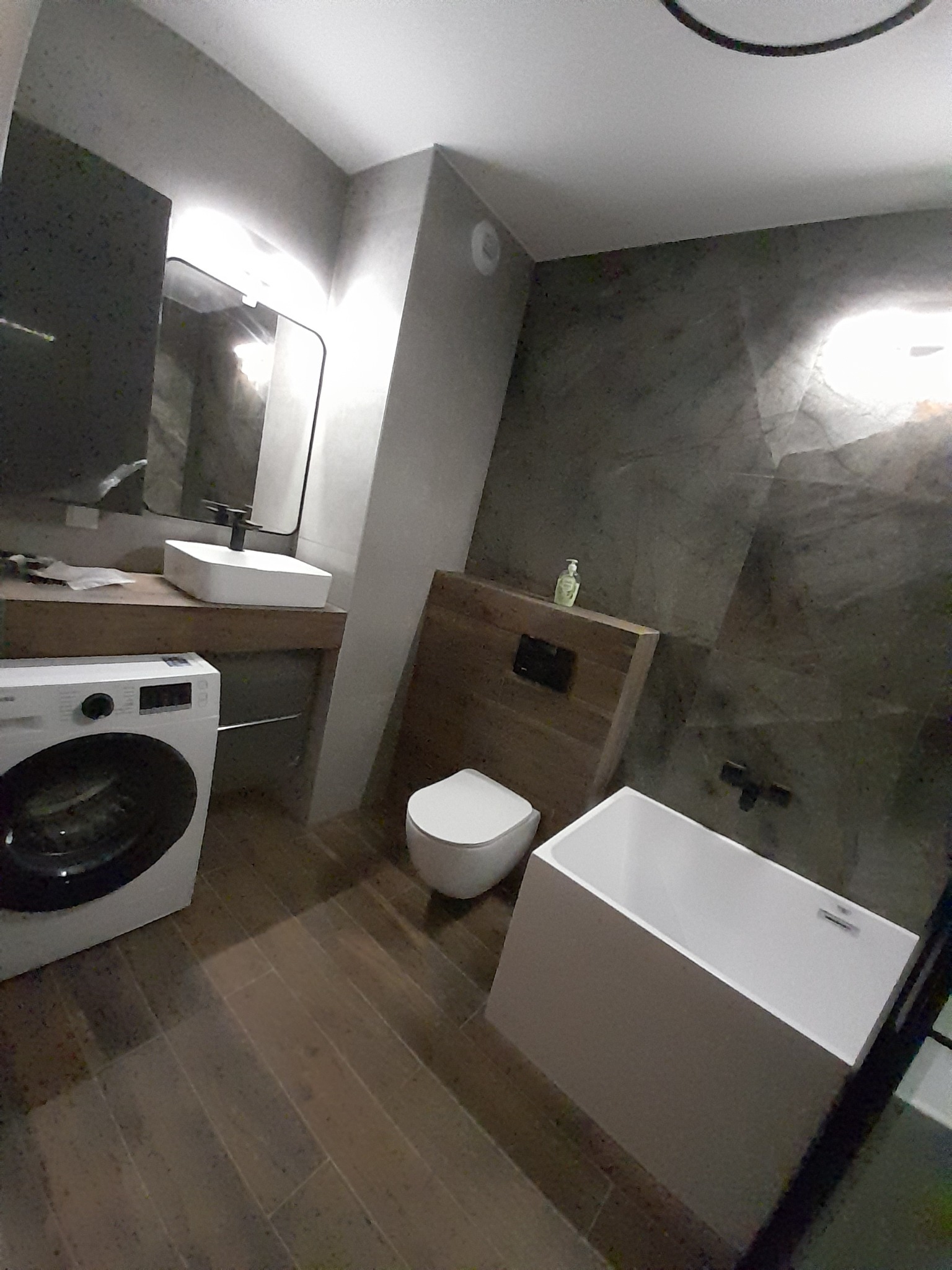 Nowoczesna łazienka z drewnianymi akcentami: umywalka nablatowa, toaleta podwieszana, wanna prostokątna i pralka automatyczna na tle szarej ściany z efektem kamienia.