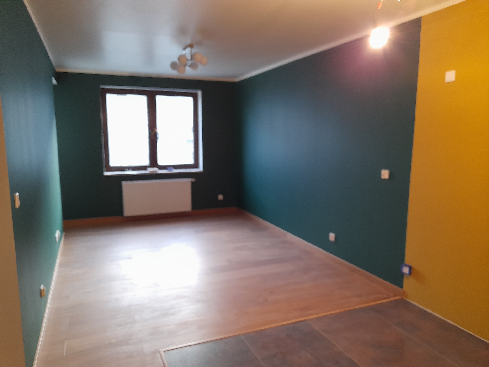 Pokój po zmianie koloru ścian na butelkową zieleń i żółty, z nową drewnianą podłogą, widoczne gniazdka elektryczne.