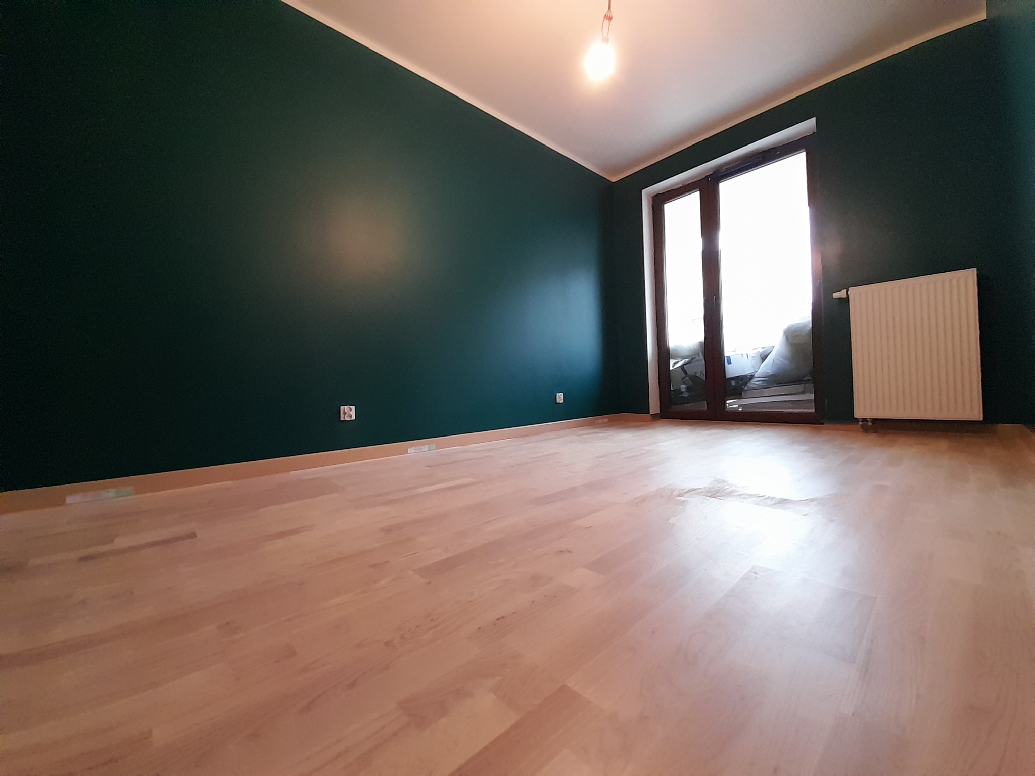 Pomalowane na ciemnozielono ściany w pustym pokoju z jasnymi panelami podłogowymi i dużym oknem balkonowym z grzejnikiem pod nim.