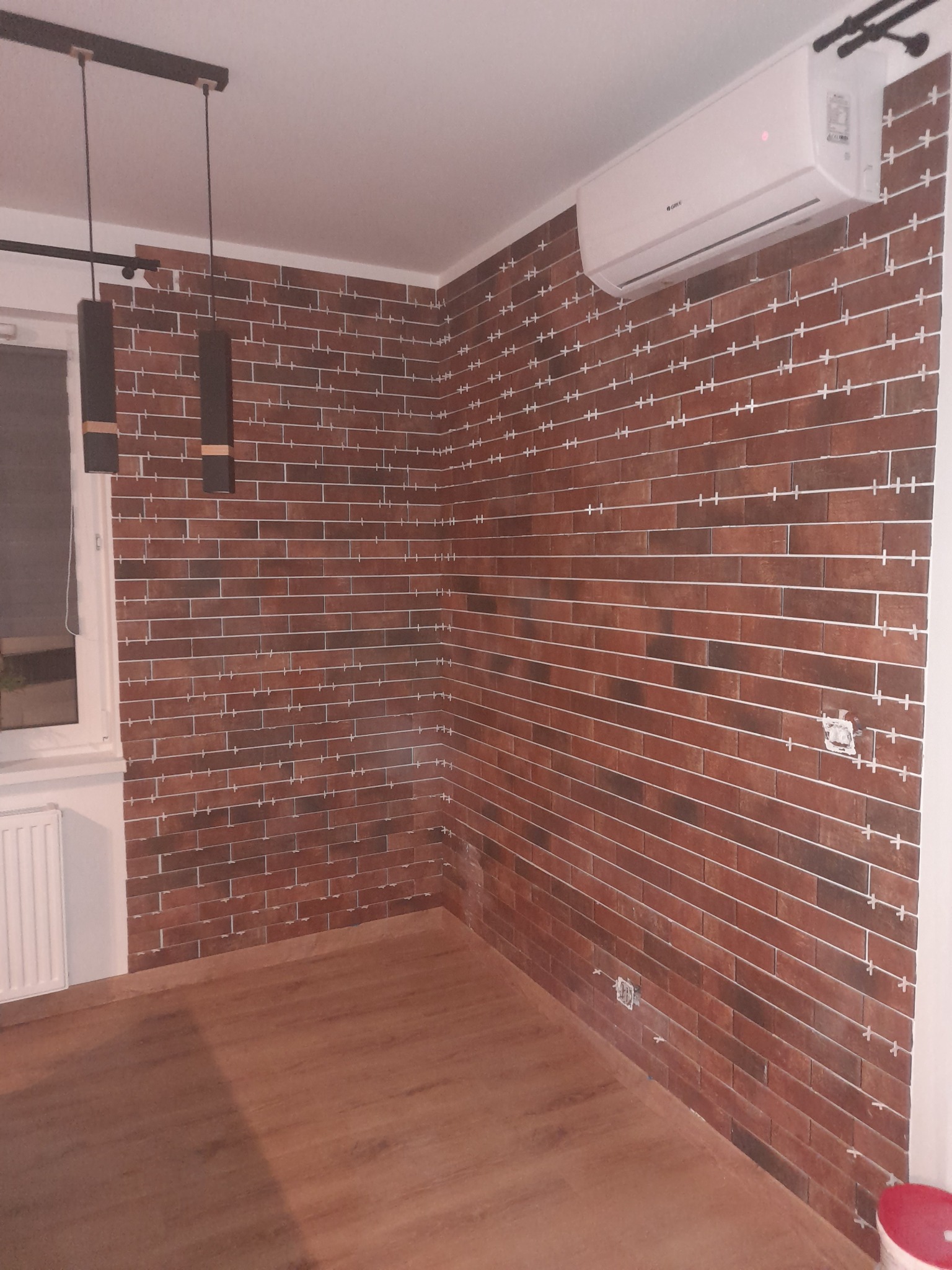 Ściana wyłożona cegiełkami klinkierowymi z krzyżykami dystansowymi, widoczny klimatyzator i lampa wisząca, świeżo położona podłoga.