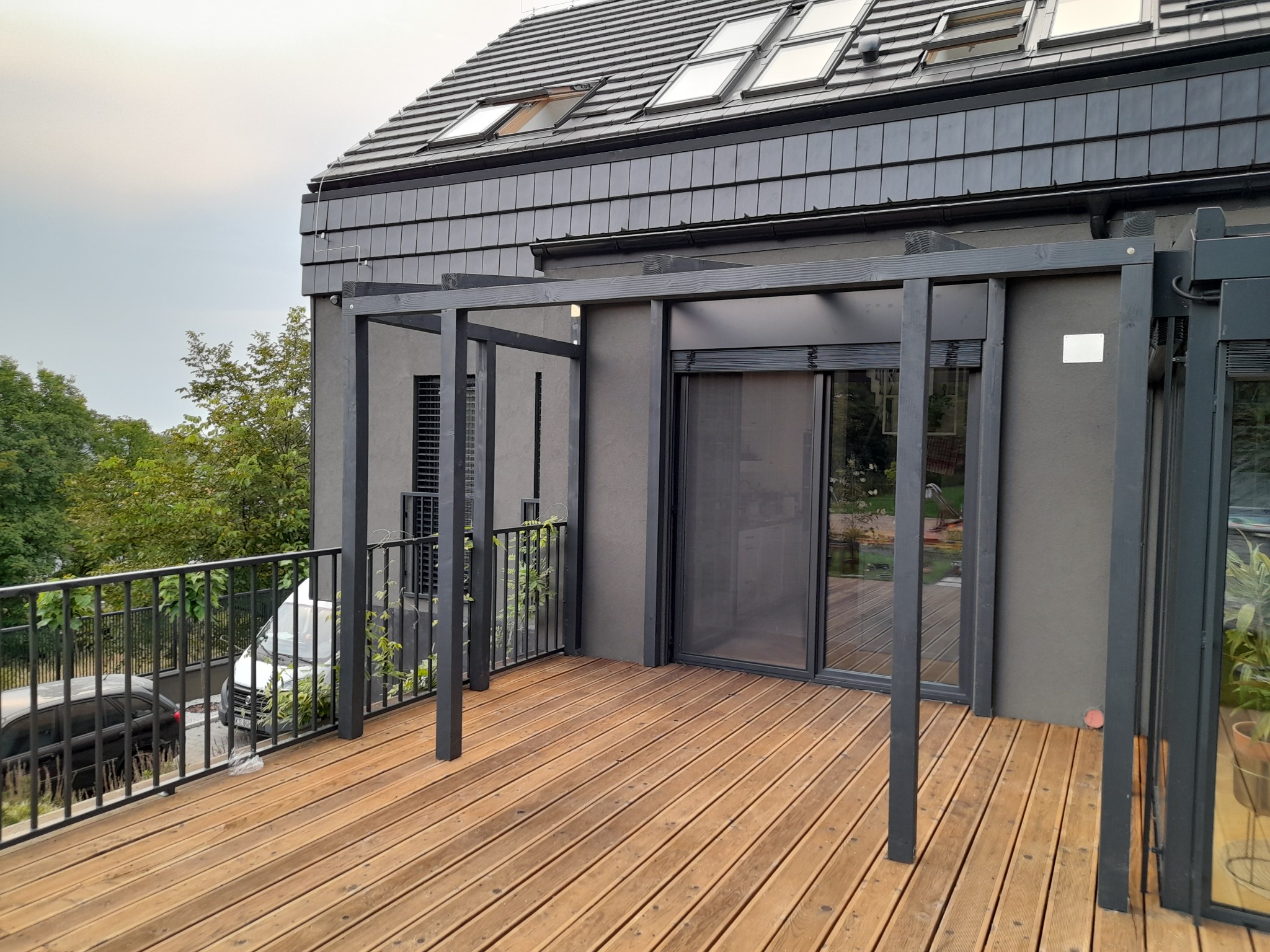 Drewniany taras z balustradą i pergolą przy nowoczesnym domu z szarą elewacją i oknami dachowymi, widok na ogród i zaparkowane samochody.