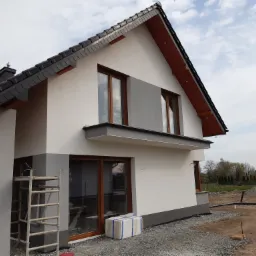 Dwukondygnacyjny dom z szarą dachówką i białymi elewacjami, drewniane okna, widoczne rusztowanie i przygotowany teren wokół budynku.