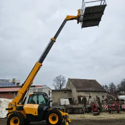 Żółto-czarna ładowarka teleskopowa JCB z podnośnikiem koszowym na tle budynków gospodarczych i pochmurnego nieba.