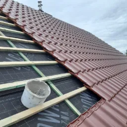 Układanie brązowej dachówki na łatach, widoczna membrana dachowa z nadrukiem, obok białe wiadro z metalowymi elementami mocującymi.