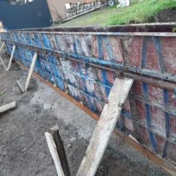 Szczegółowe ujęcie szalunku fundamentowego z czerwono-niebieskimi panelami, wzmocnionego metalowymi rurami i drewnianymi podporami na placu budowy.