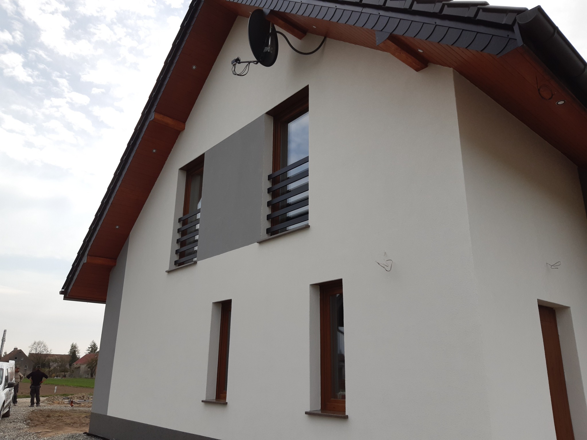 Wykończony dom murowany z jasną elewacją, drewnianymi oknami i ciemnym dachem, widoczna antena satelitarna na szczycie ściany.