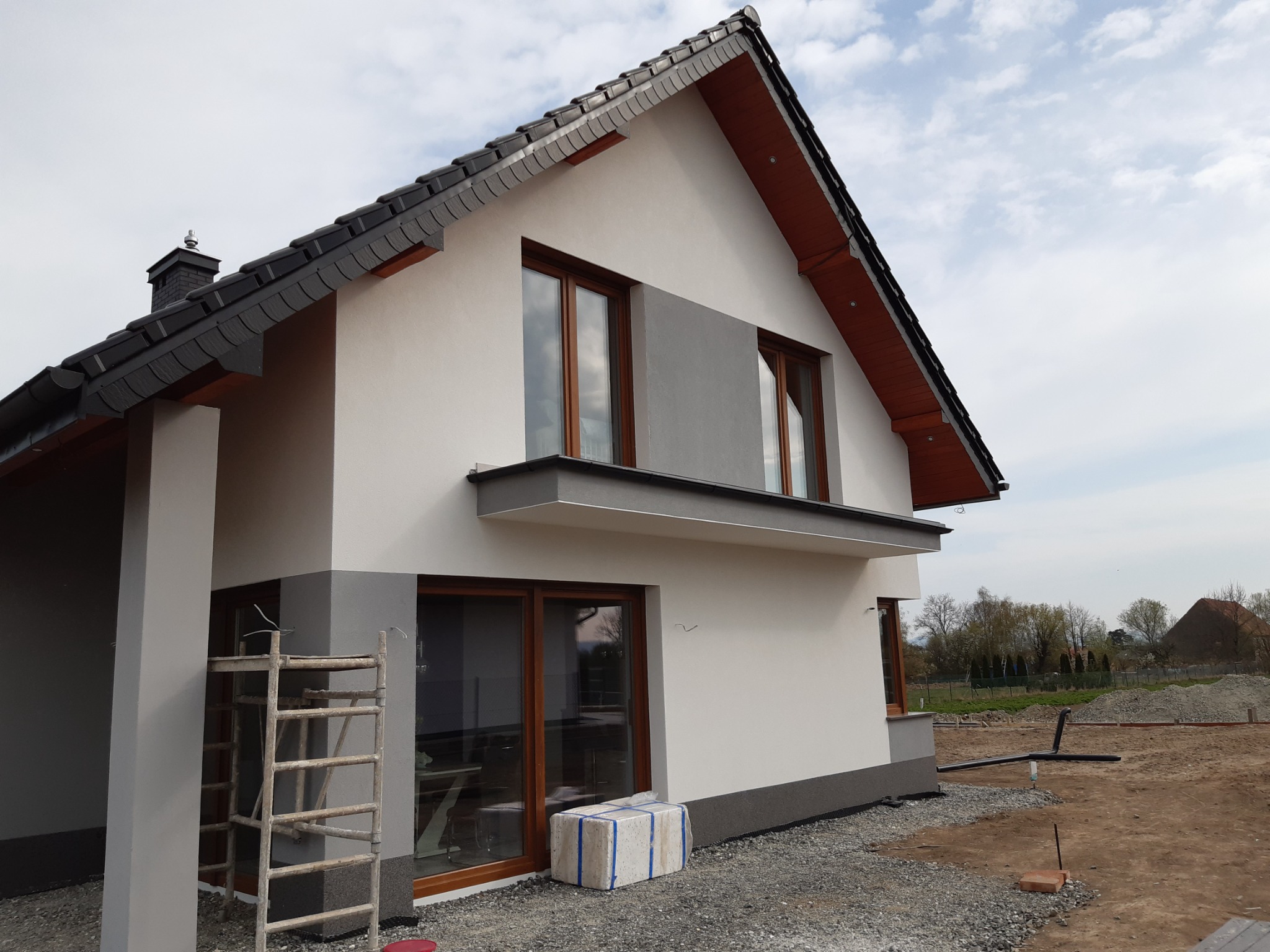 Dwukondygnacyjny dom z szarą dachówką i białymi elewacjami, drewniane okna, widoczne rusztowanie i przygotowany teren wokół budynku.