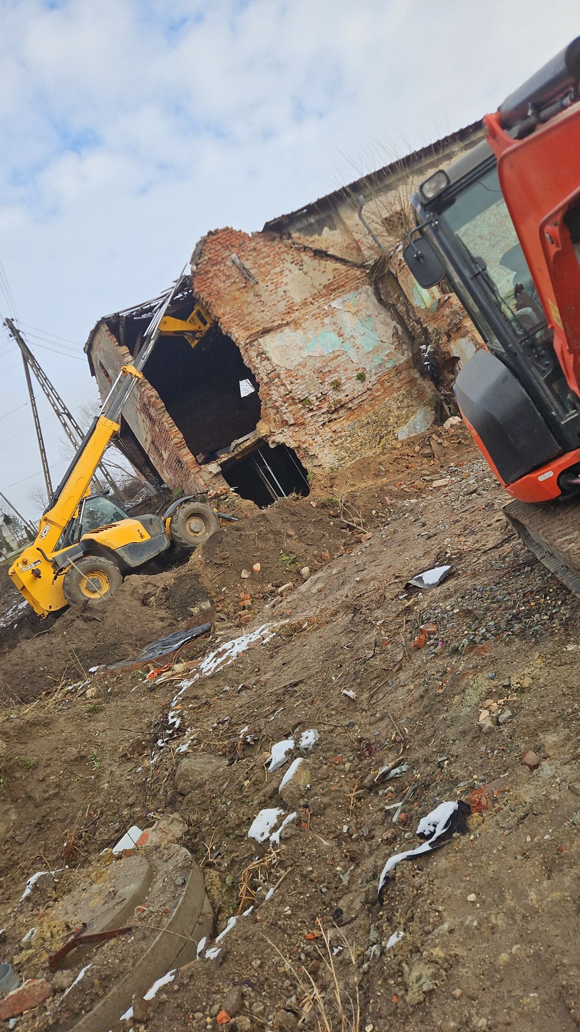 Żółta ładowarka teleskopowa i czerwona koparka na zboczu ziemi przed częściowo zburzonym, ceglanym budynkiem z widocznymi otworami okiennymi i resztkami tynku.