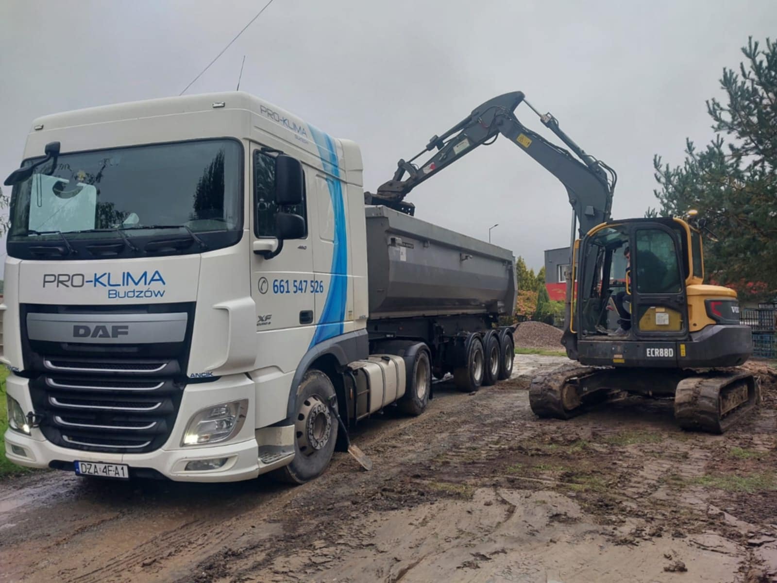 Żółta koparka ładuje gruz do skrzyni ciężarówki firmy PRO-KLIMA na błotnistym terenie.