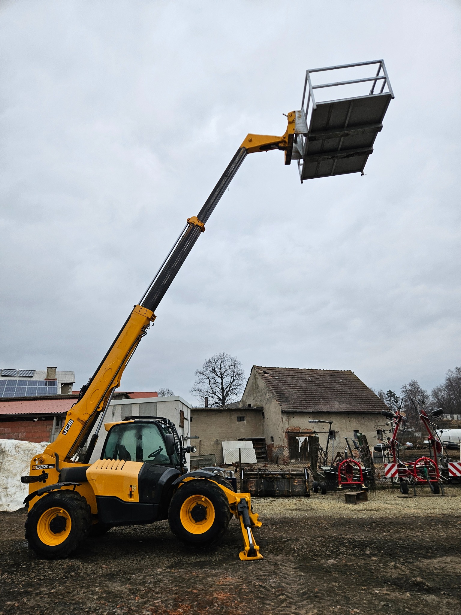 Żółto-czarna ładowarka teleskopowa JCB z podnośnikiem koszowym na tle budynków gospodarczych i pochmurnego nieba.