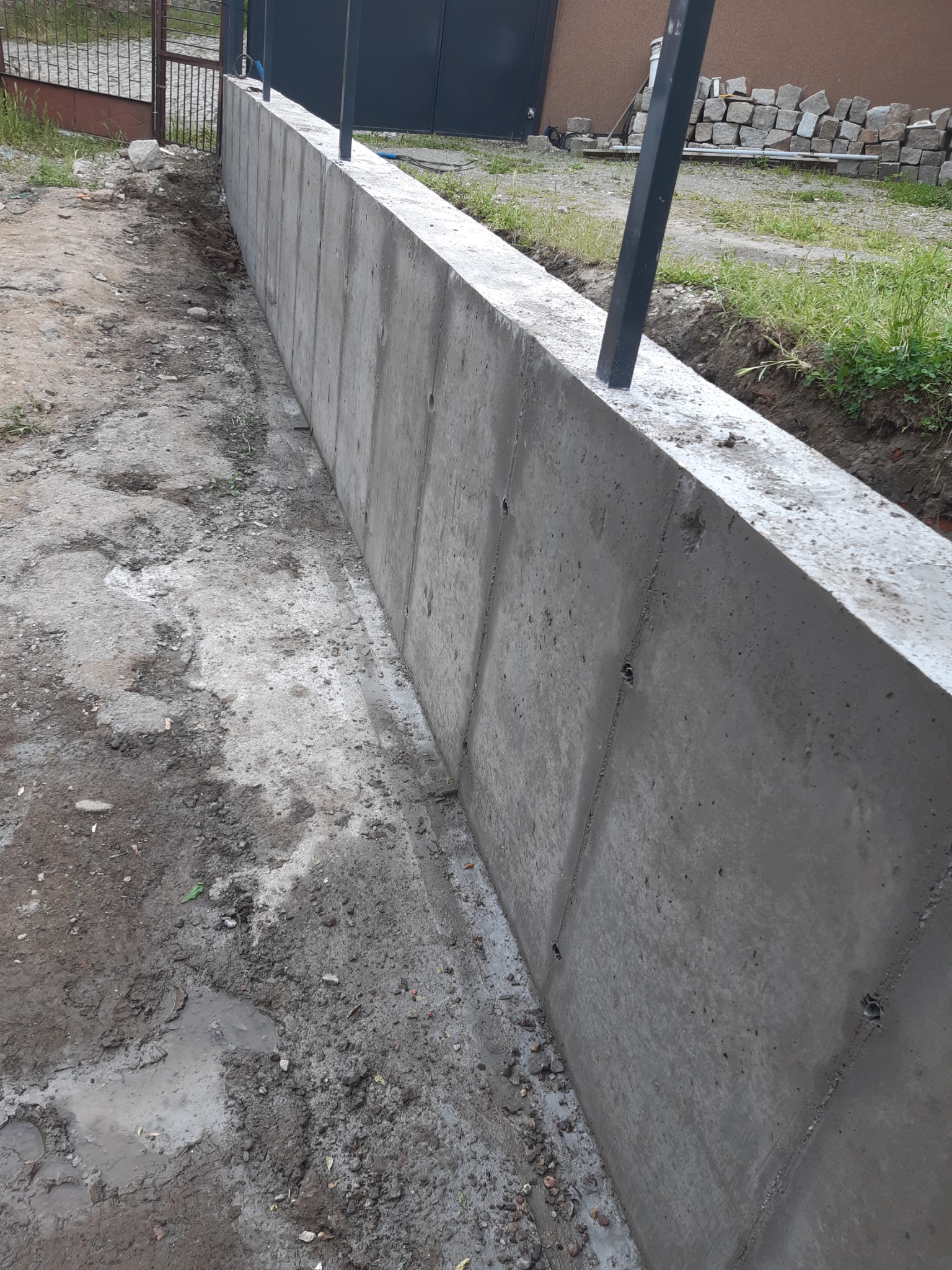 Betonowy murek oporowy w trakcie budowy, widoczne ślady po szalunkach, obok nierówny teren z błotem i stertą kamieni.