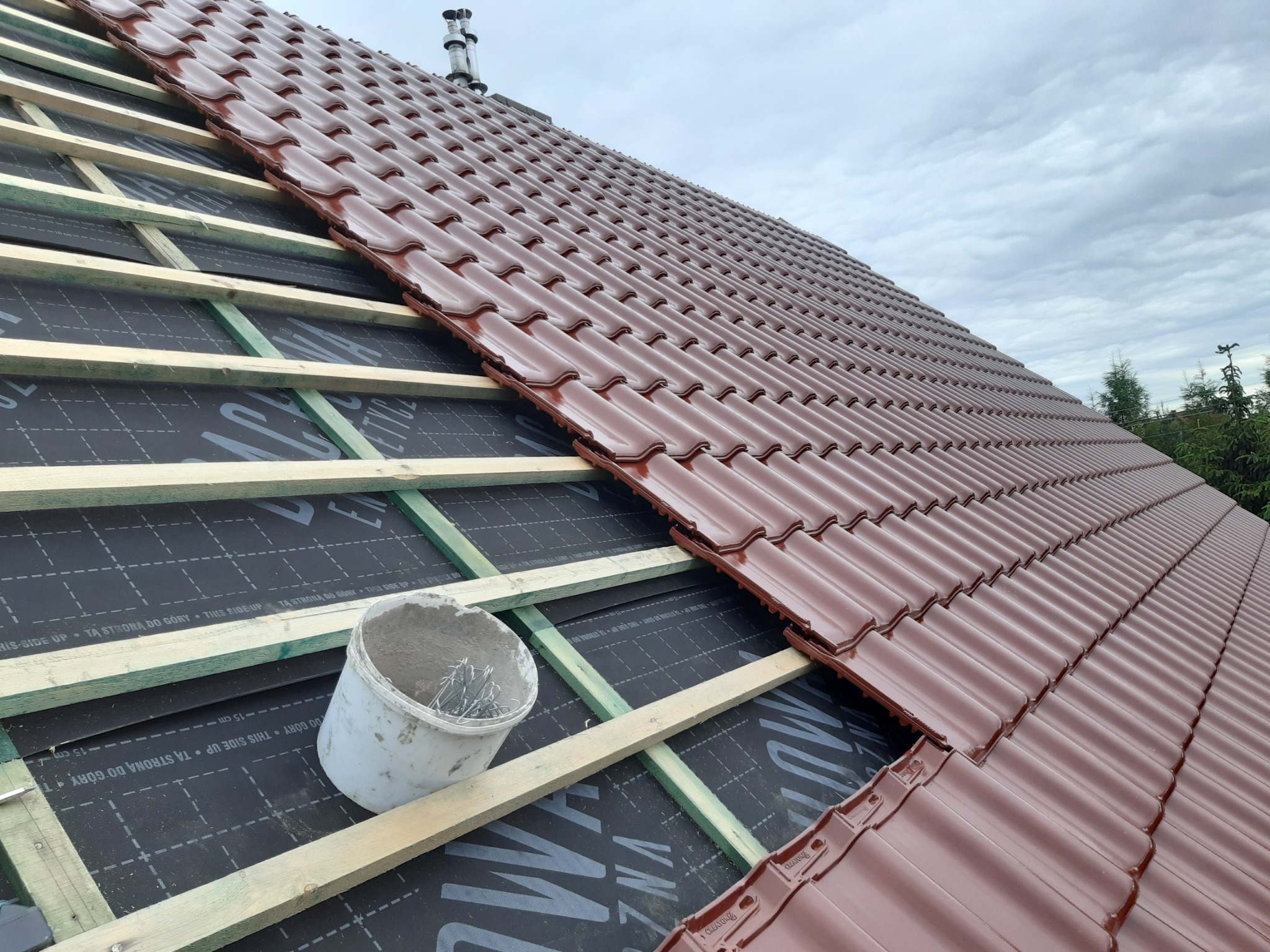Układanie brązowej dachówki na łatach, widoczna membrana dachowa z nadrukiem, obok białe wiadro z metalowymi elementami mocującymi.