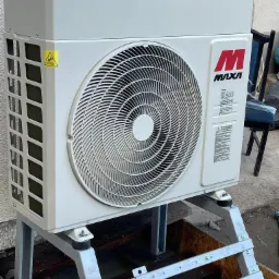 Zewnętrzna jednostka pompy ciepła marki MVXA zamontowana na metalowej konstrukcji wsporczej, widoczne narzędzia na górnej obudowie, żółta naklejka ostrzegawcza na boku.