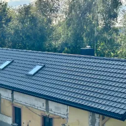 Nowy, grafitowy dach z dwoma oknami połaciowymi na domu w trakcie rozbudowy, widok z góry na tle zieleni. Widoczne surowe ściany i fragment zadaszenia.