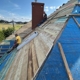 Kosar-bud - Montaż drewnianej konstrukcji dachu z kominem w słoneczny dzień. Widoczni dekarze podczas pracy, niebieska membrana dachowa i drewniane deski na tle błękitnego nieba.