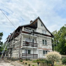 Kosar-bud - Dwupiętrowy dom w trakcie rozbudowy, otoczony rusztowaniem. Widoczni pracownicy na drabinie, częściowo wykończona elewacja i elementy dachu.