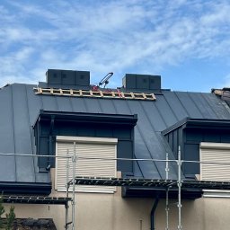 Kosar-bud - Szary dach z blachy na domu z lukarnami i rusztowaniem. Na dachu widoczne kominy wentylacyjne i drabina dekarska. Dodatkowo widoczne solary na sąsiednim dachu.