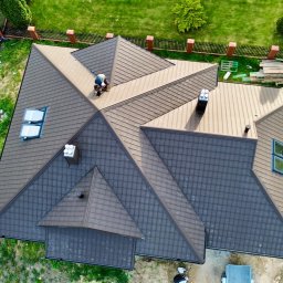 Kosar-bud - Widok z góry na brązowy dach wielospadowy z blachodachówki, z kominami i oknami dachowymi. Na dachu pracuje dekarz. Wokół domu zieleń i ogrodzenie.