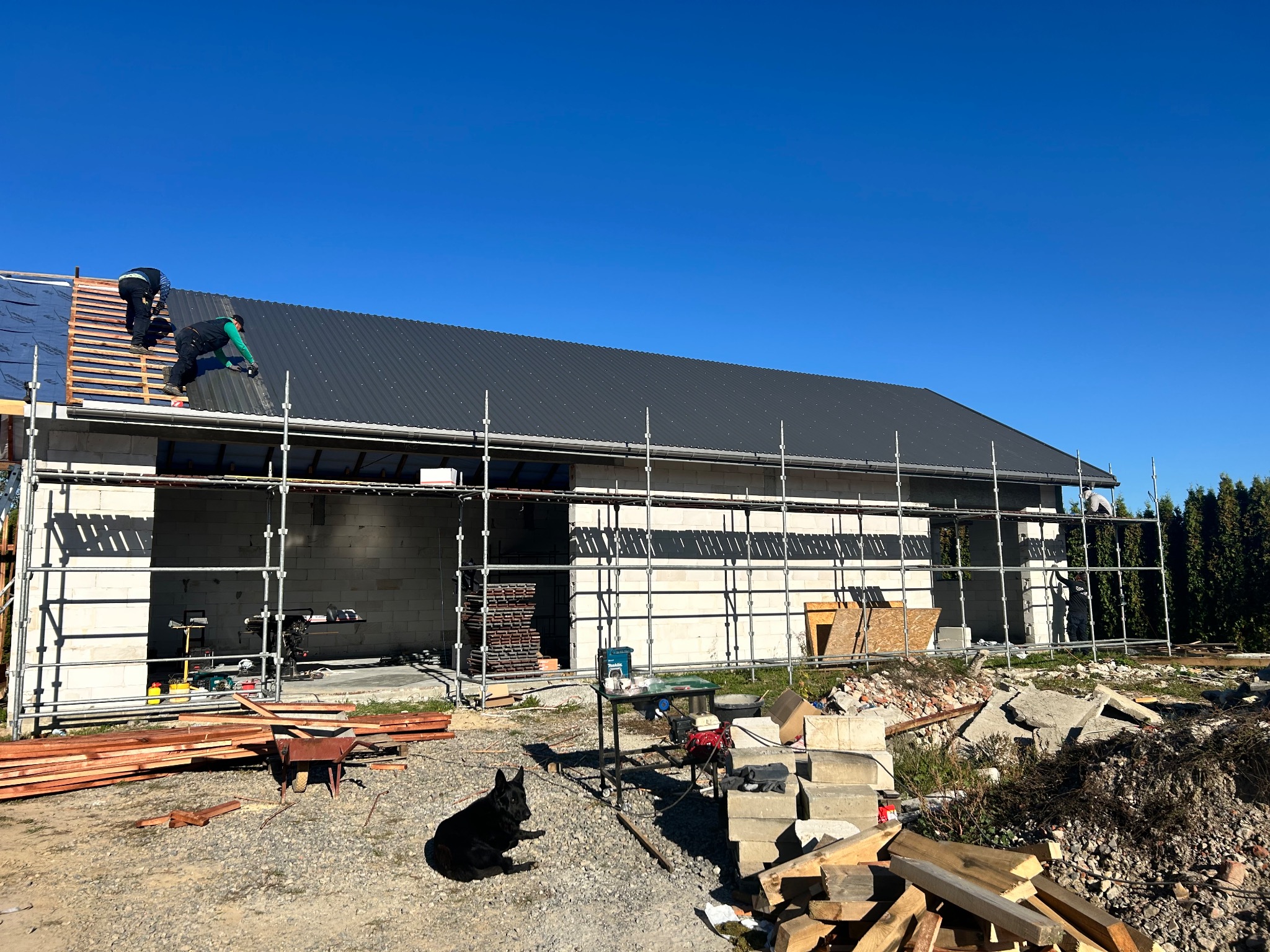 Montaż ciemnoszarej blachy na dachu budynku w trakcie budowy. Widoczne rusztowania, narzędzia i czarny pies leżący na ziemi. Prace dekarskie w słoneczny dzień.