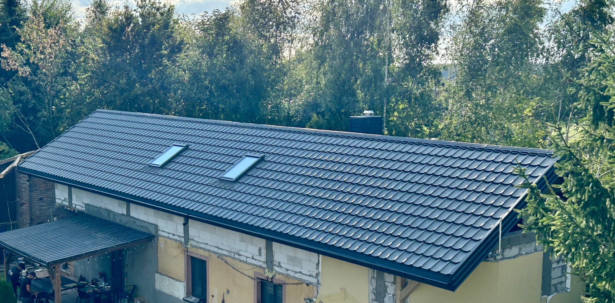 Nowy, grafitowy dach z dwoma oknami połaciowymi na domu w trakcie rozbudowy, widok z góry na tle zieleni. Widoczne surowe ściany i fragment zadaszenia.