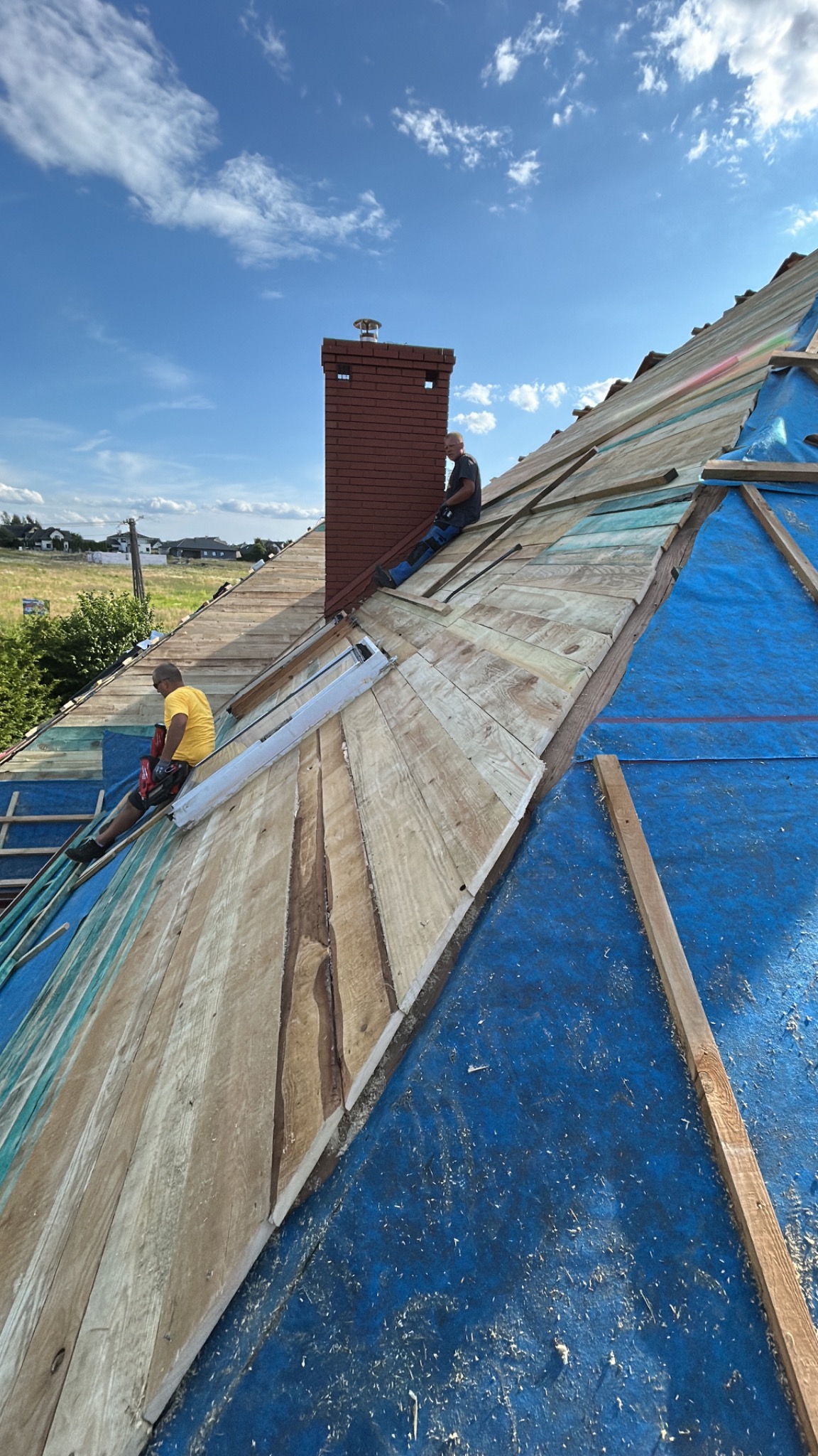 Montaż drewnianej konstrukcji dachu z kominem w słoneczny dzień. Widoczni dekarze podczas pracy, niebieska membrana dachowa i drewniane deski na tle błękitnego nieba.