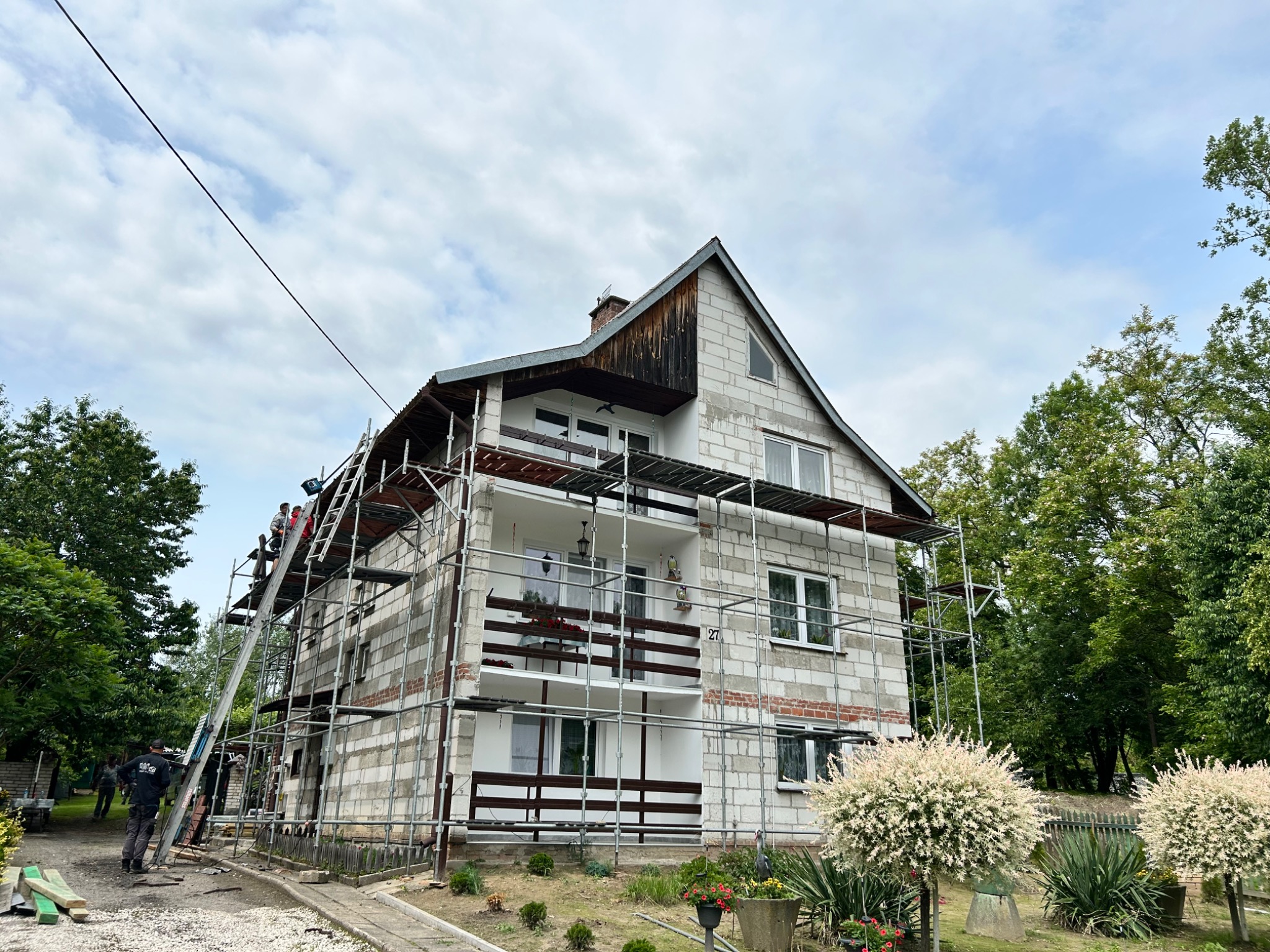 Dwupiętrowy dom w trakcie rozbudowy, otoczony rusztowaniem. Widoczni pracownicy na drabinie, częściowo wykończona elewacja i elementy dachu.