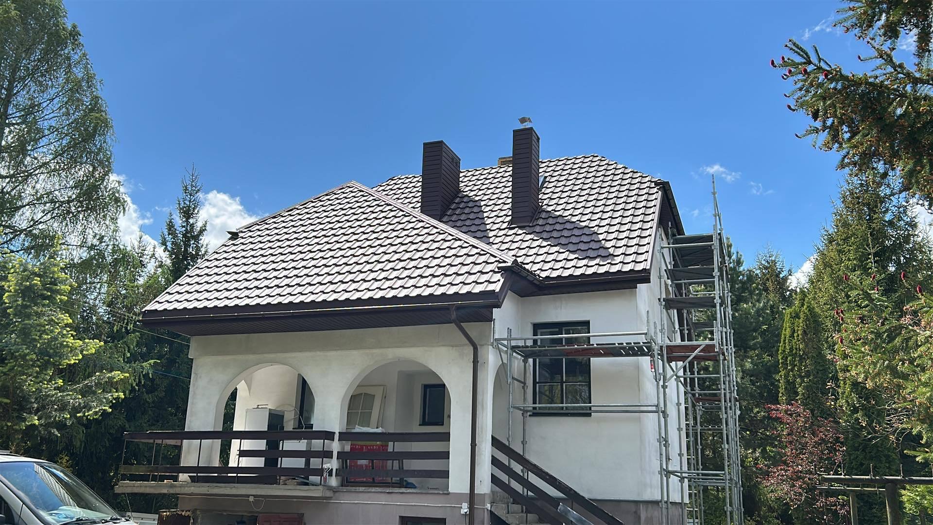 Biały dom z brązowym dachem z dachówki, dwa kominy, rusztowanie przy ścianie, błękitne niebo w tle.
