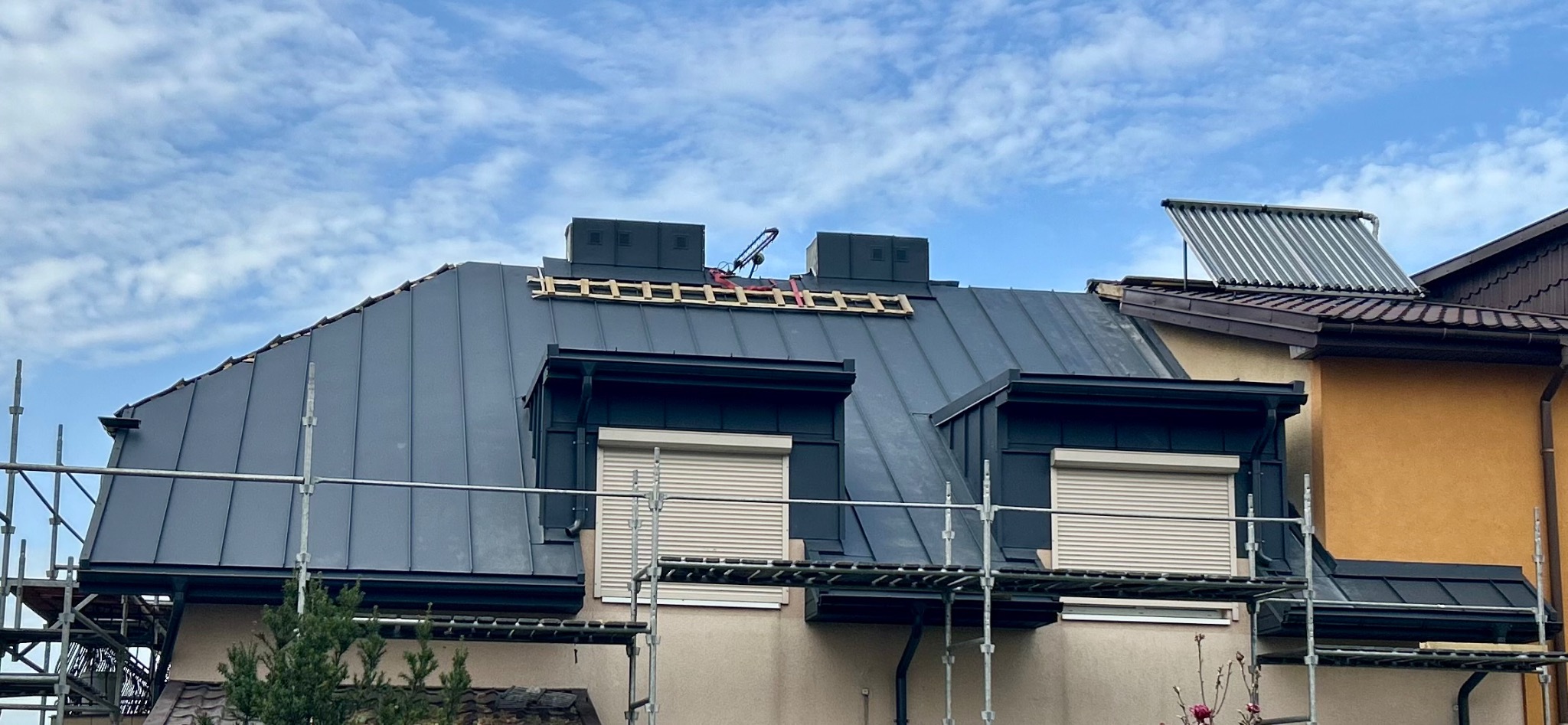 Szary dach z blachy na domu z lukarnami i rusztowaniem. Na dachu widoczne kominy wentylacyjne i drabina dekarska. Dodatkowo widoczne solary na sąsiednim dachu.