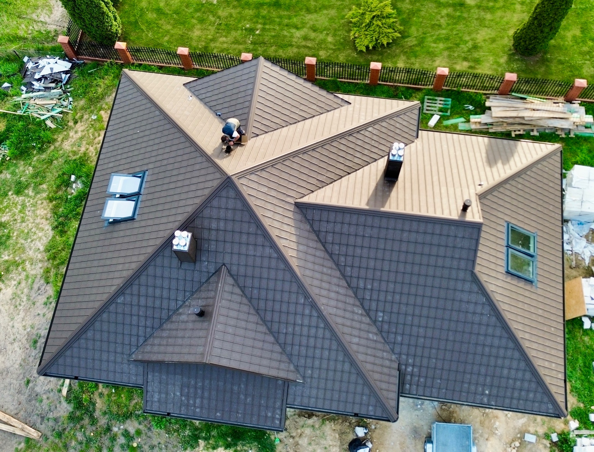 Widok z góry na brązowy dach wielospadowy z blachodachówki, z kominami i oknami dachowymi. Na dachu pracuje dekarz. Wokół domu zieleń i ogrodzenie.