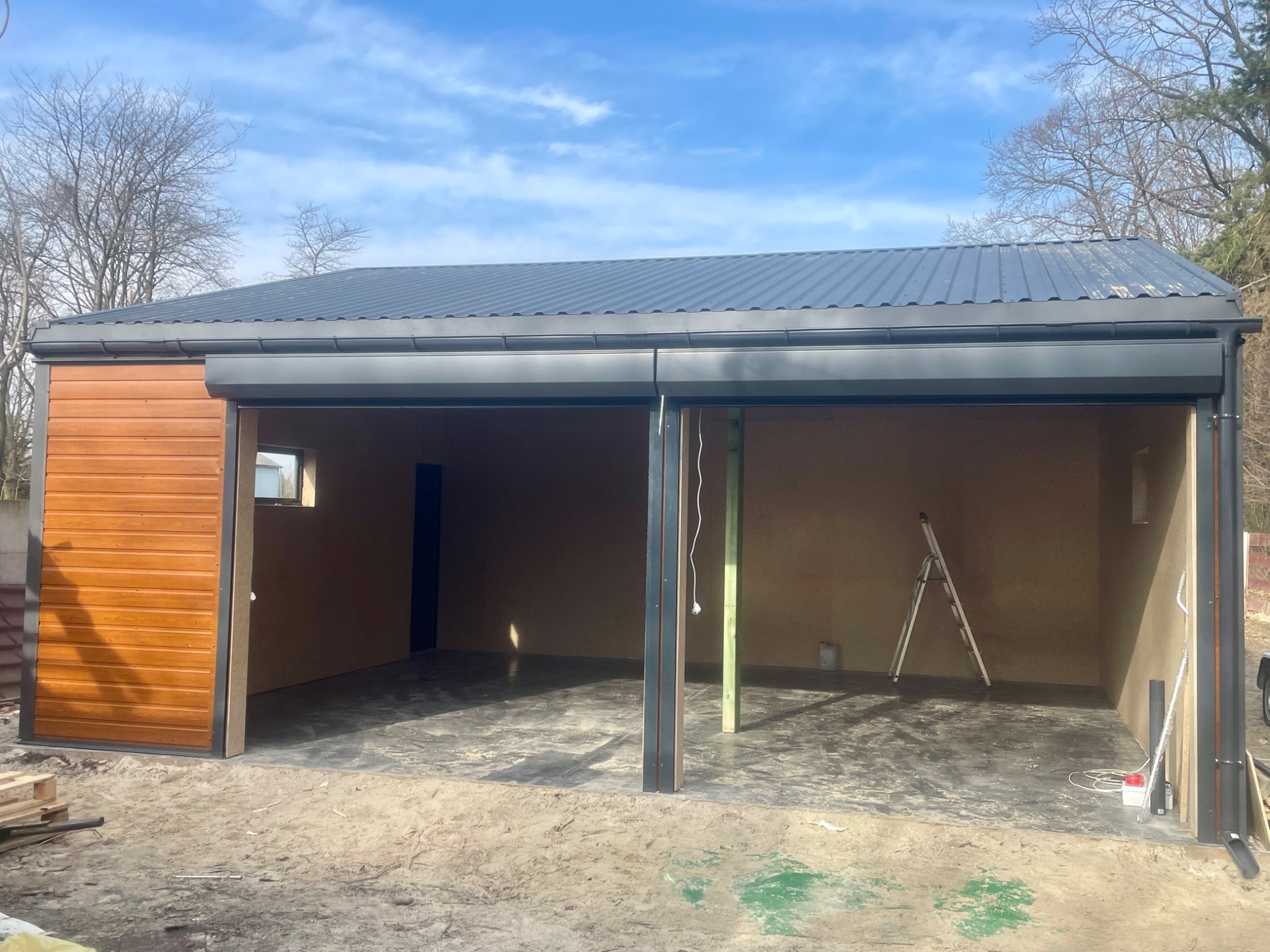 Nowoczesny garaż blaszany z drewnianą elewacją i rolowanymi bramami, w trakcie budowy. Widoczna drabina wewnątrz, błękitne niebo w tle.