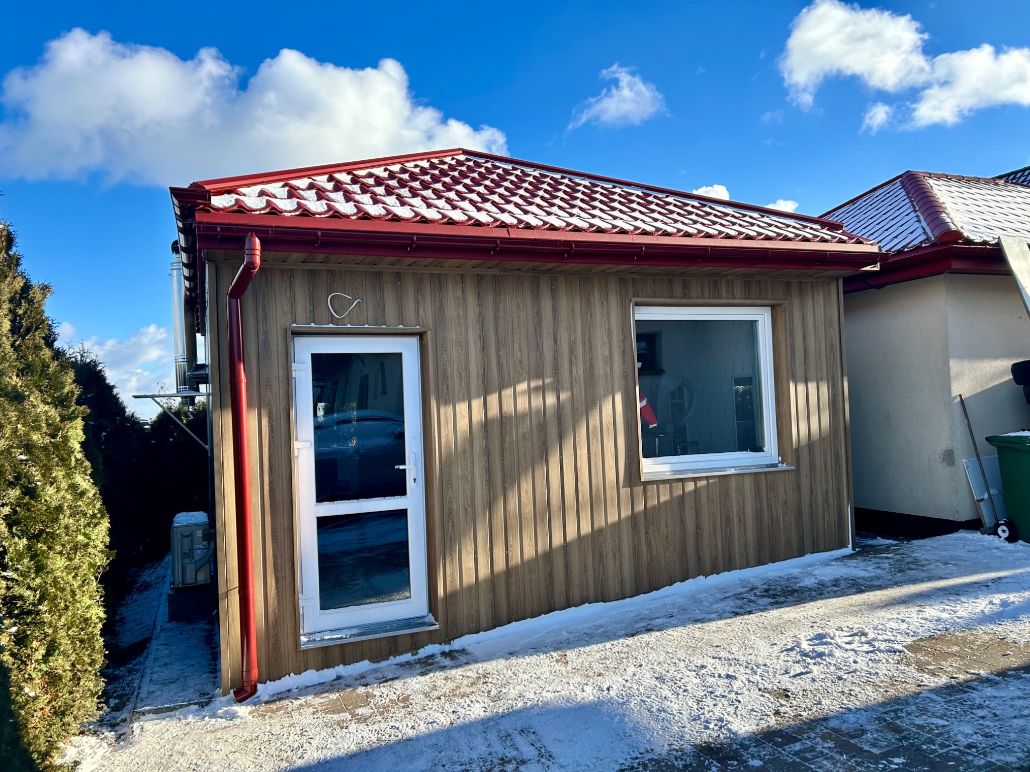 Nowoczesny, drewniany budynek z czerwoną dachówką ceramiczną pokrytą śniegiem, widoczny w zimowej scenerii. Elewacja pionowa, okno PCV, rynna w kolorze dachu.