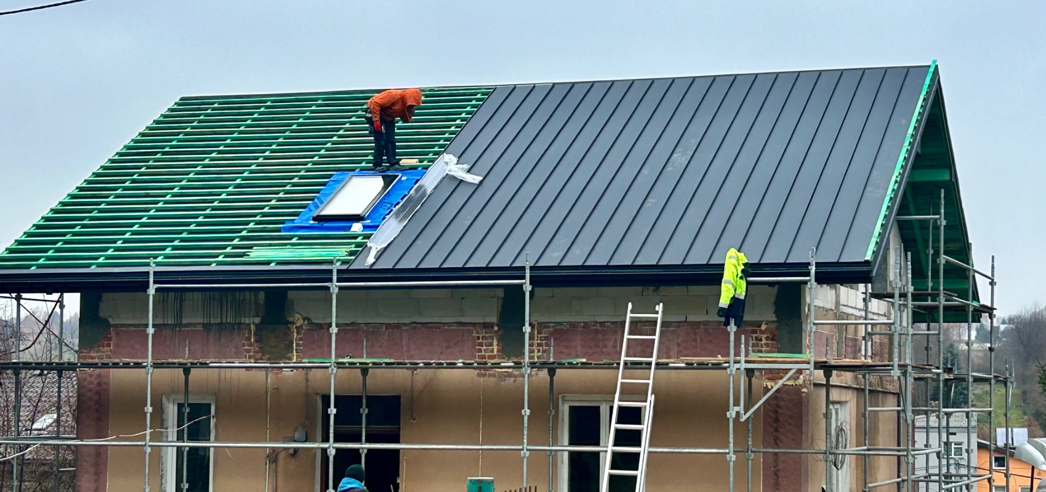 Montaż dachu z blachy na budynku w trakcie budowy. Widoczne rusztowania, więźba dachowa i pracownik w pomarańczowej kurtce. Część dachu pokryta blachą, część z zieloną konstrukcją.