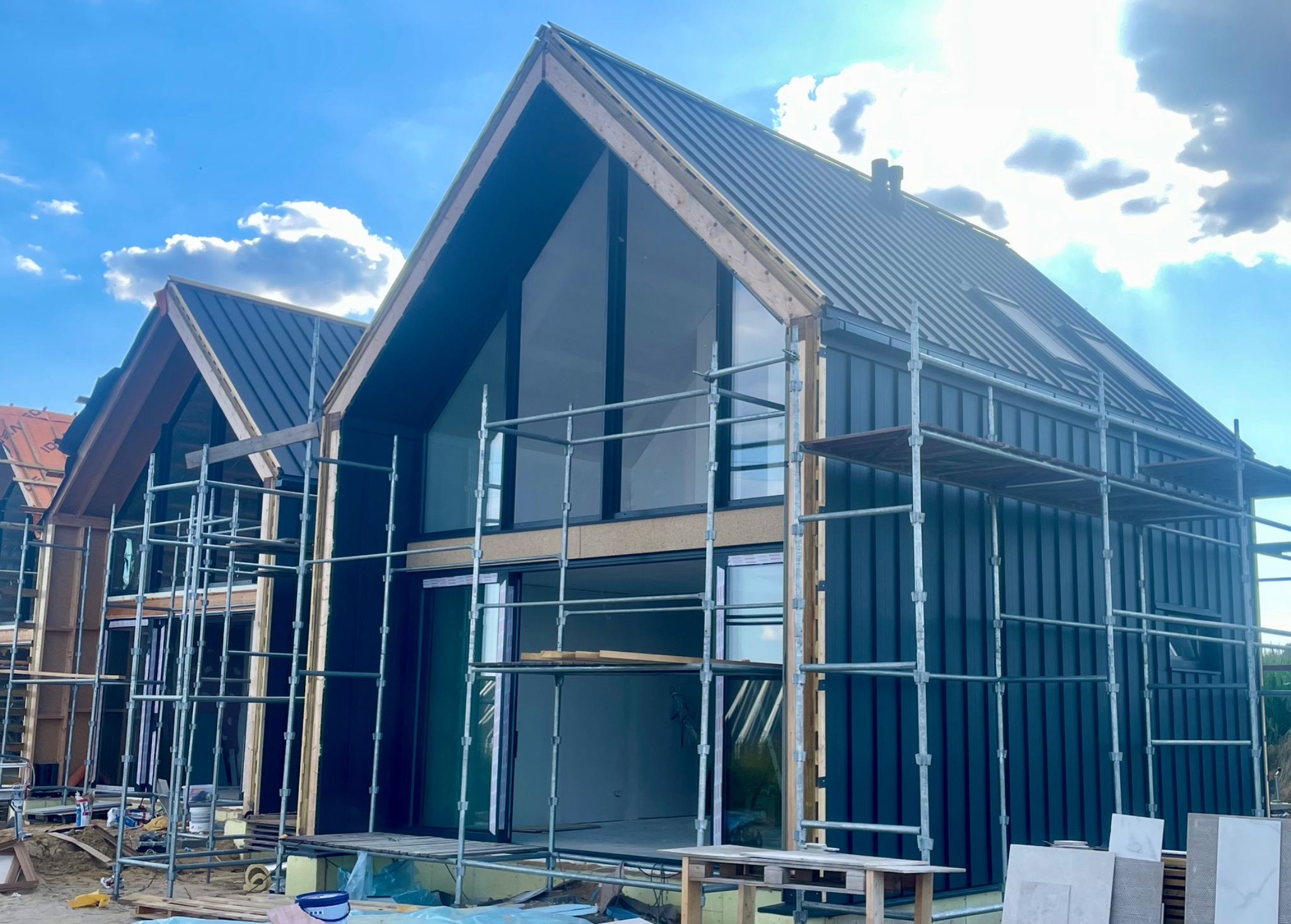 Wznoszenie nowoczesnego domu z czarną blachą trapezową na dachu. Widoczne rusztowania i duże okna. Budowa w toku, jasne niebo w tle.