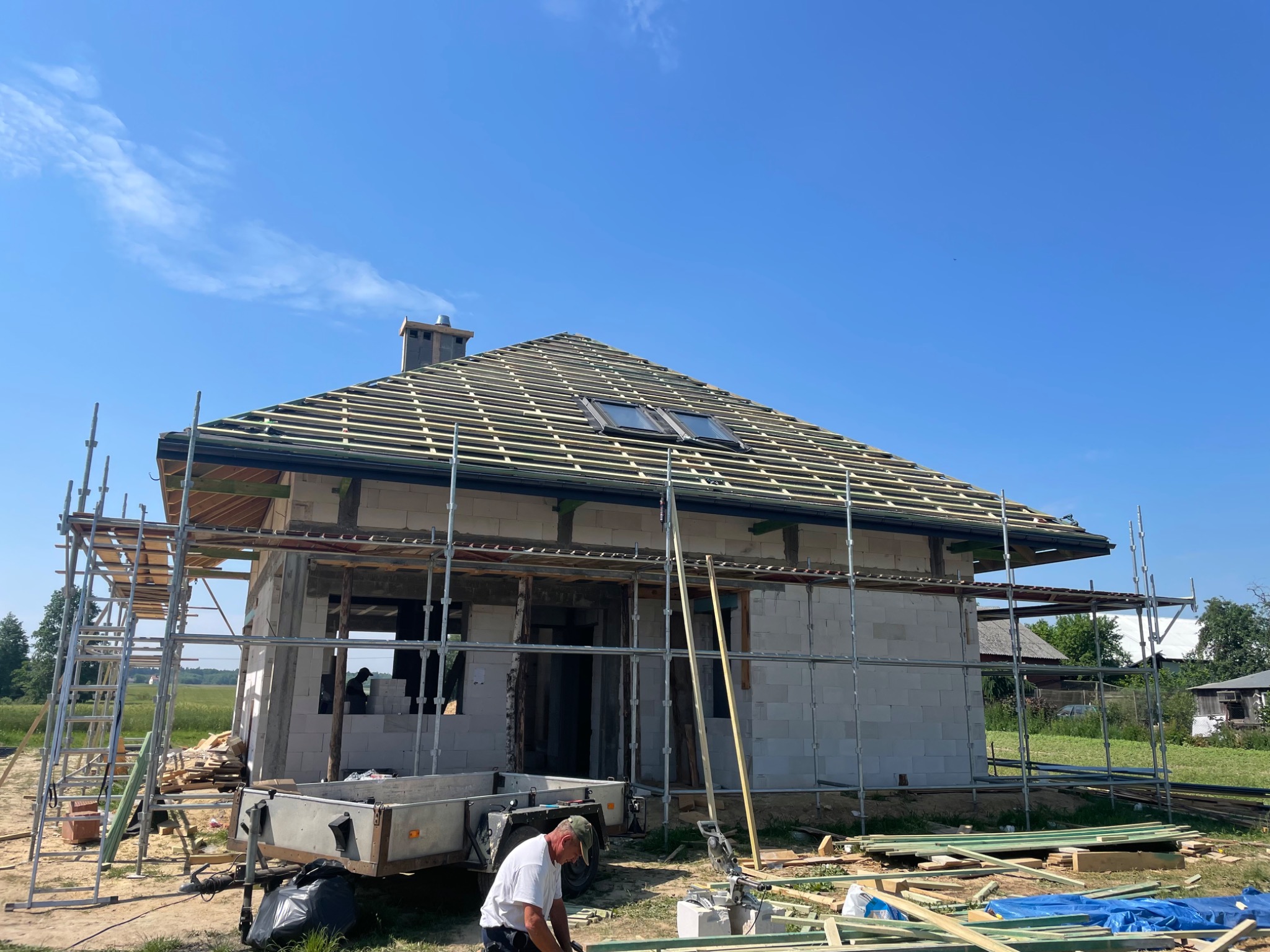 Budowa domu jednorodzinnego w trakcie montażu więźby dachowej z widocznymi oknami dachowymi, rusztowaniami i pracującym mężczyzną przy przyczepce z narzędziami.