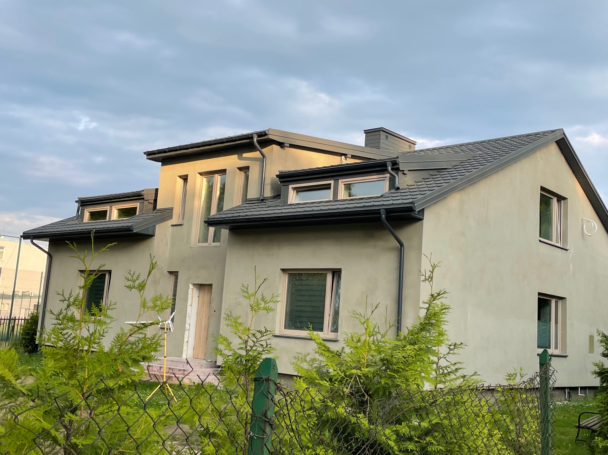 Nowoczesny dom jednorodzinny z szarą blachodachówką płaską, lukarnami i świeżo pomalowaną elewacją w odcieniach beżu, z rynnami w kolorze dachu, widziany zza ogrodzenia z krzewami.
