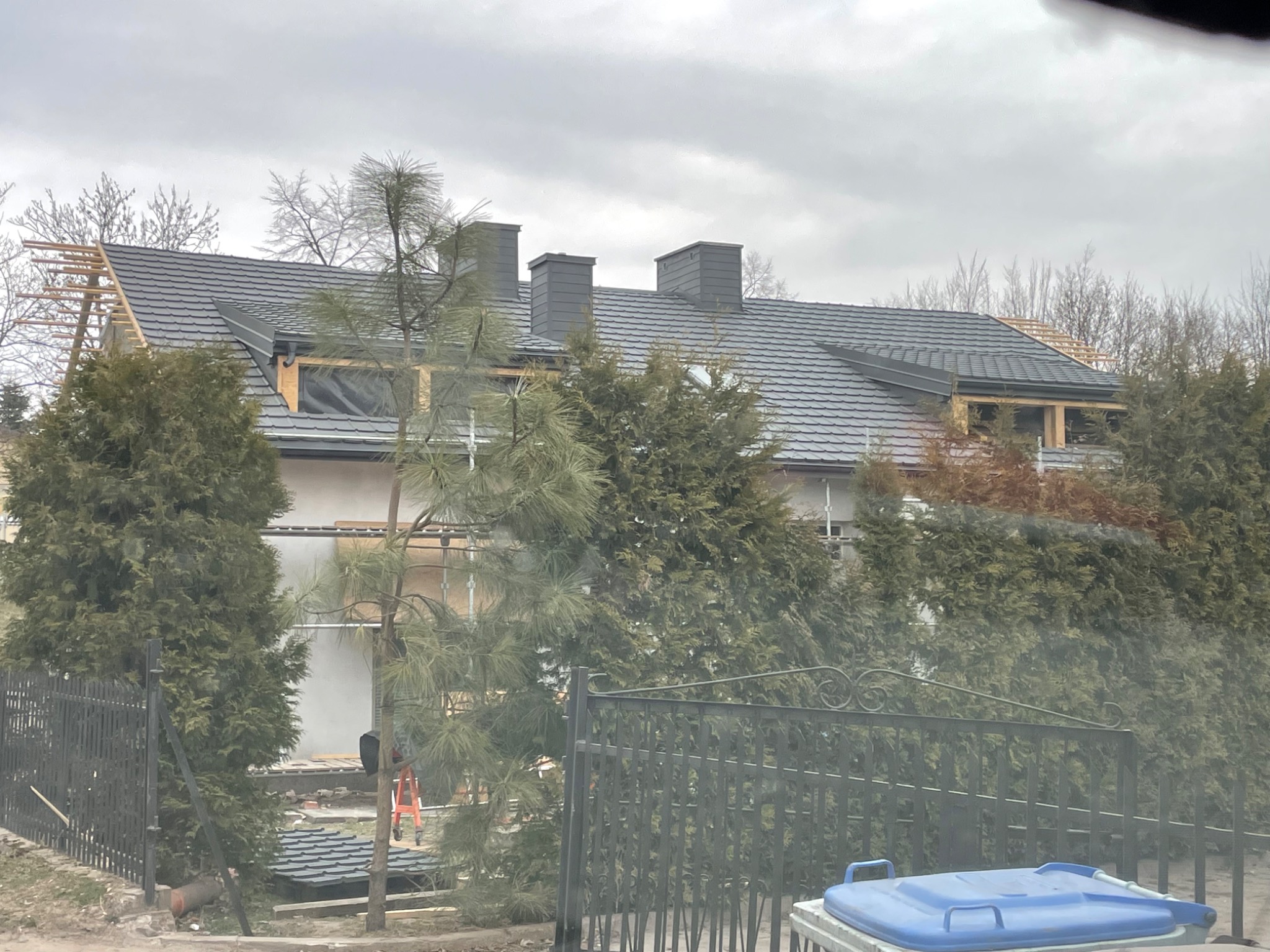 Dom w trakcie prac dekarskich, widoczne elementy więźby dachowej i fragment pokrycia dachowego w kolorze grafitowym, kominy obłożone blachą. Na pierwszym planie zieleń i fragment ogrodzenia.