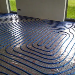 Niebieskie rury ogrzewania podłogowego ułożone spiralnie na folii z czerwoną siatką w jasnym, surowym pomieszczeniu z dużym oknem.