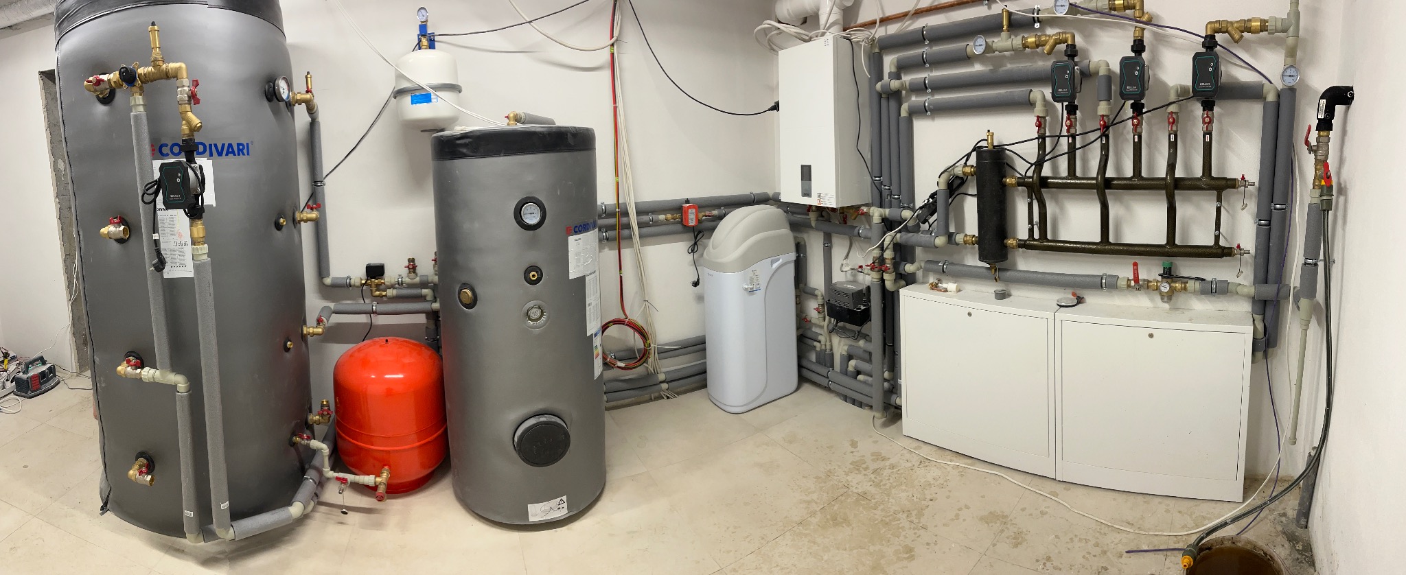 Szeroki kadr na kotłownię z dwoma szarymi zbiornikami Cordivari, czerwonym zbiornikiem przeponowym, zmiękczaczem wody, rozdzielaczem grzewczym i białą szafką, w tle instalacja grzewcza z rurami...