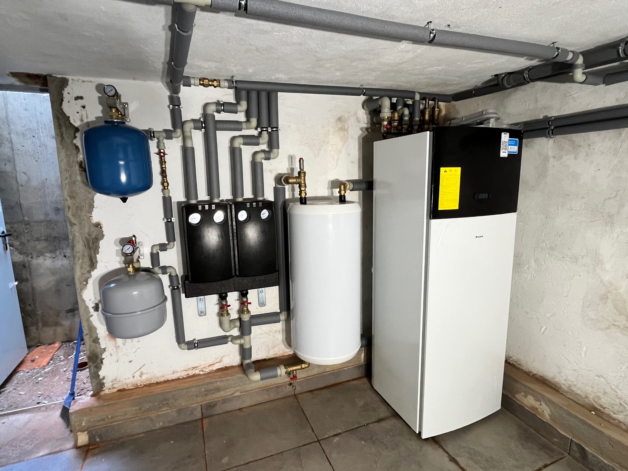 Biała pompa ciepła Daikin z podłączonymi zbiornikami wyrównawczymi i zasobnikiem ciepłej wody w kotłowni z surowymi ścianami i szarymi rurami instalacyjnymi.