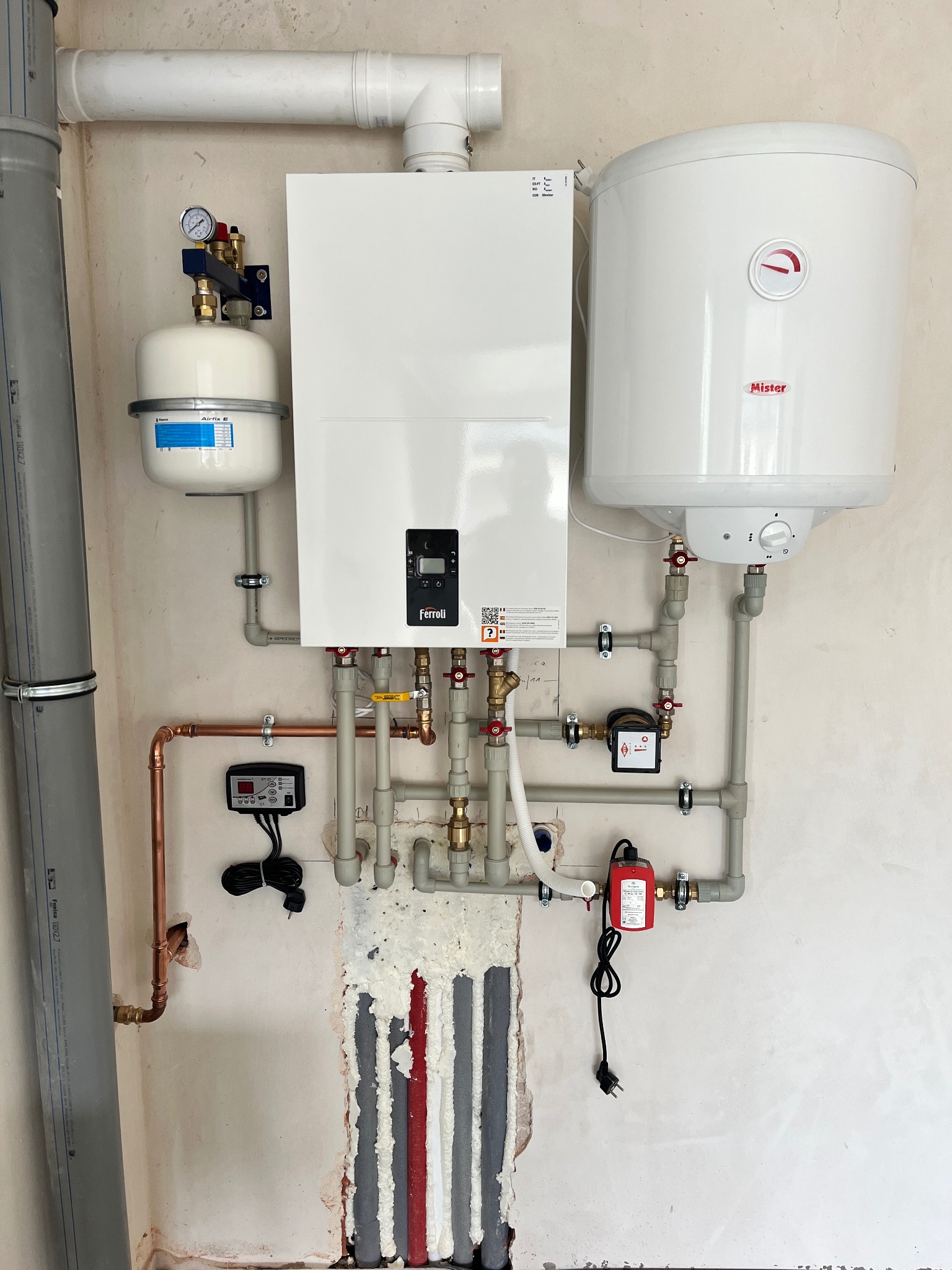 Biały piec centralnego ogrzewania marki Ferroli z podłączonym zasobnikiem Mister, widoczne rury miedziane i plastikowe, manometr, zawory, sterownik oraz izolowane rury w ścianie w trakcie montażu.