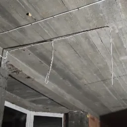 Betonowy sufit z widocznymi przewodami elektrycznymi, częściowo zamocowanymi, z fragmentami zwisającymi. W tle widoczne okno i surowa ściana z cegły.