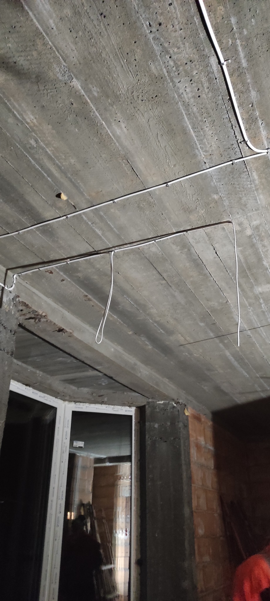 Betonowy sufit z widocznymi przewodami elektrycznymi, częściowo zamocowanymi, z fragmentami zwisającymi. W tle widoczne okno i surowa ściana z cegły.