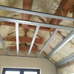 Poddasze ocieplone pianą poliuretanową, widoczne drewniane krokwie i metalowy stelaż pod zabudowę, fragment okna w dolnej części kadru.