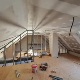 Adaptacja poddasza: metalowa konstrukcja szkieletowa pod płyty, częściowo wyszpachlowany sufit skośny z widocznymi łączeniami, podłoga z płyt OSB, wiadro z masą szpachlową i narzędzia.