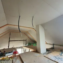 Poddasze w trakcie wykańczania płytami kartonowo-gipsowymi, widoczne drewniane elementy konstrukcyjne, izolacja, pion wentylacyjny oraz przewody elektryczne wystające ze ściany i sufitu.