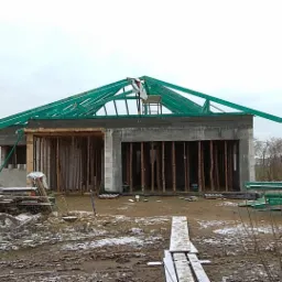 Surowy stan domu w trakcie budowy, widoczne ściany z pustaków, drewniane szalunki okienne i zielona konstrukcja dachu, na której stoi pracownik w białej odzieży.