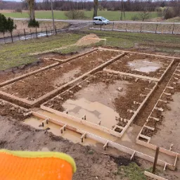 Przygotowanie terenu pod wylanie fundamentów: wykopane rowy z drewnianym szalunkiem, częściowo wypełnione błotnistą wodą, w tle hałda piasku i zaparkowany samochód.