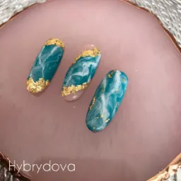 Trzy sztuczne paznokcie z turkusowo-białym marmurkowym wzorem i złotymi płatkami nałożone na różową, okrągłą podstawkę.