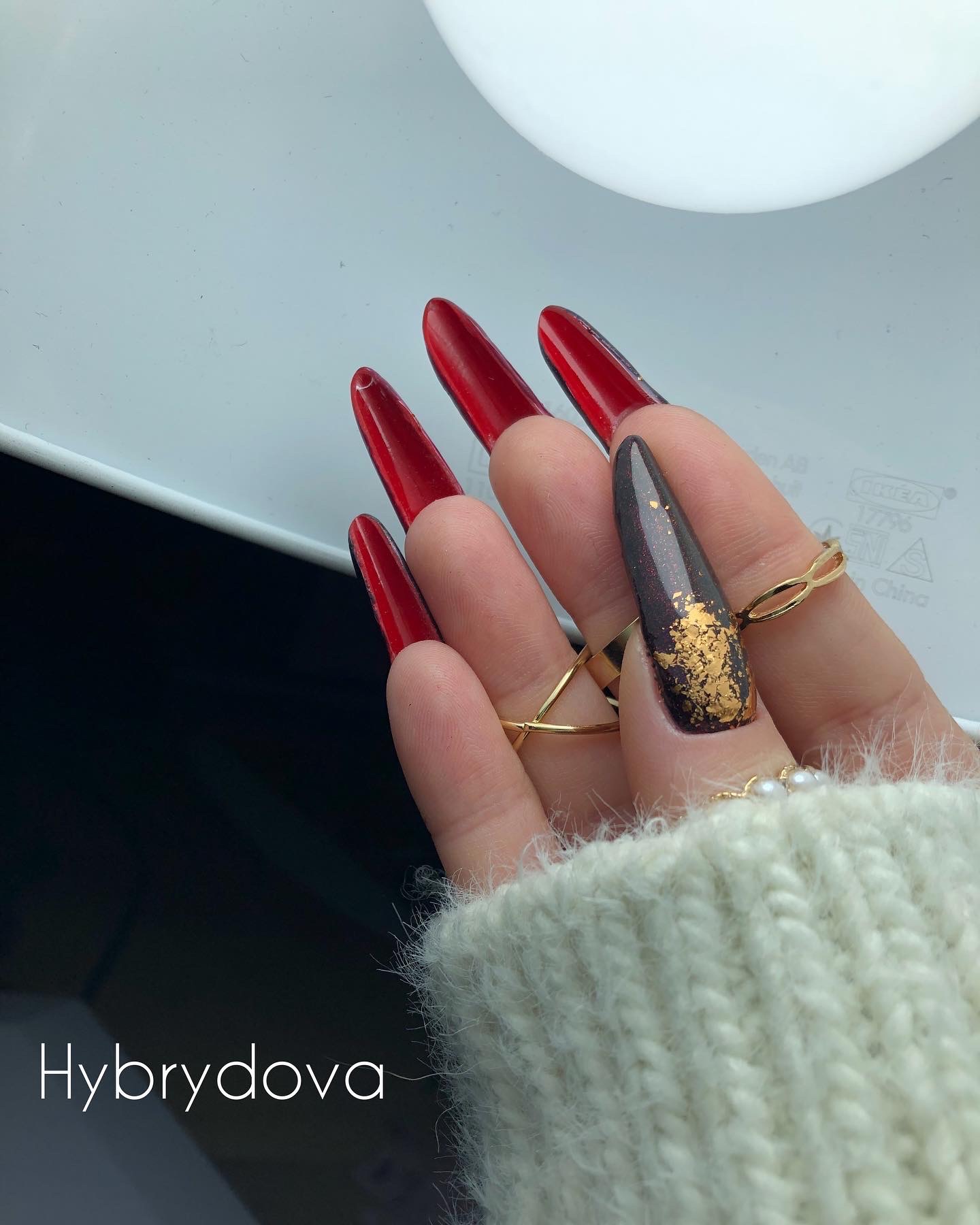 Dłoń z długimi, czerwonymi paznokciami w szpic i jednym ciemnym paznokciem z efektem złota, widoczne złote pierścionki na palcach, w tle biała powierzchnia.