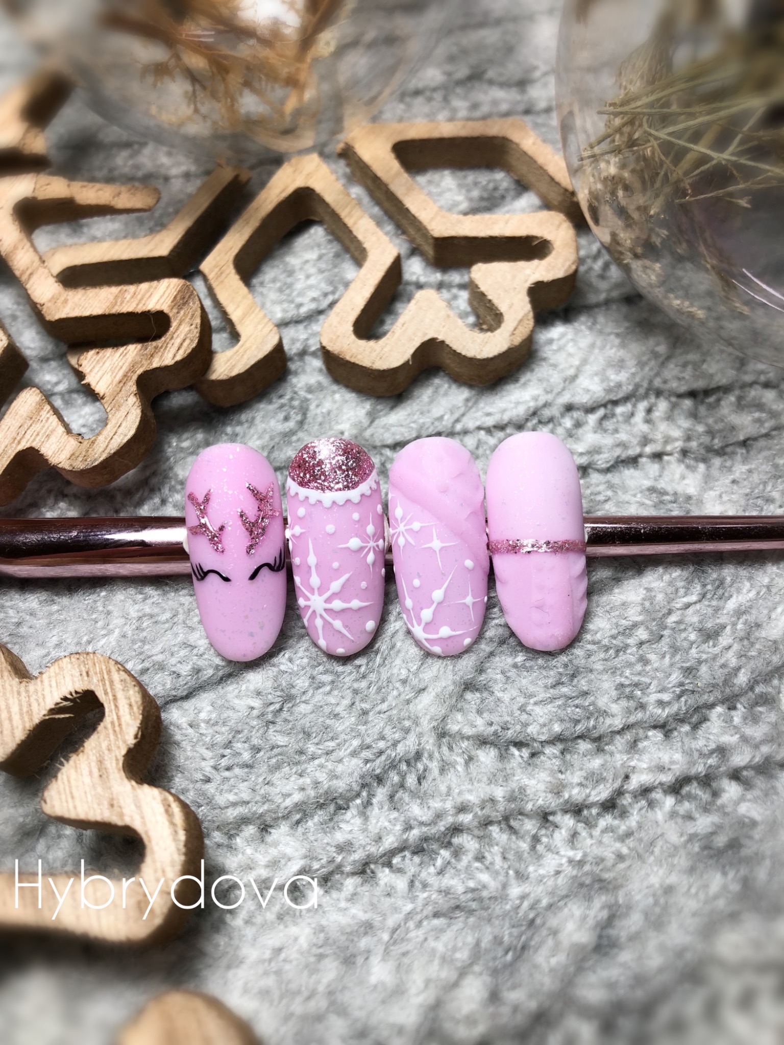 Różowe paznokcie z zimowymi zdobieniami: renifer z brokatowymi rogami, śnieżynki i brokatowy pasek, ułożone na szarym swetrze z drewnianymi ozdobami.